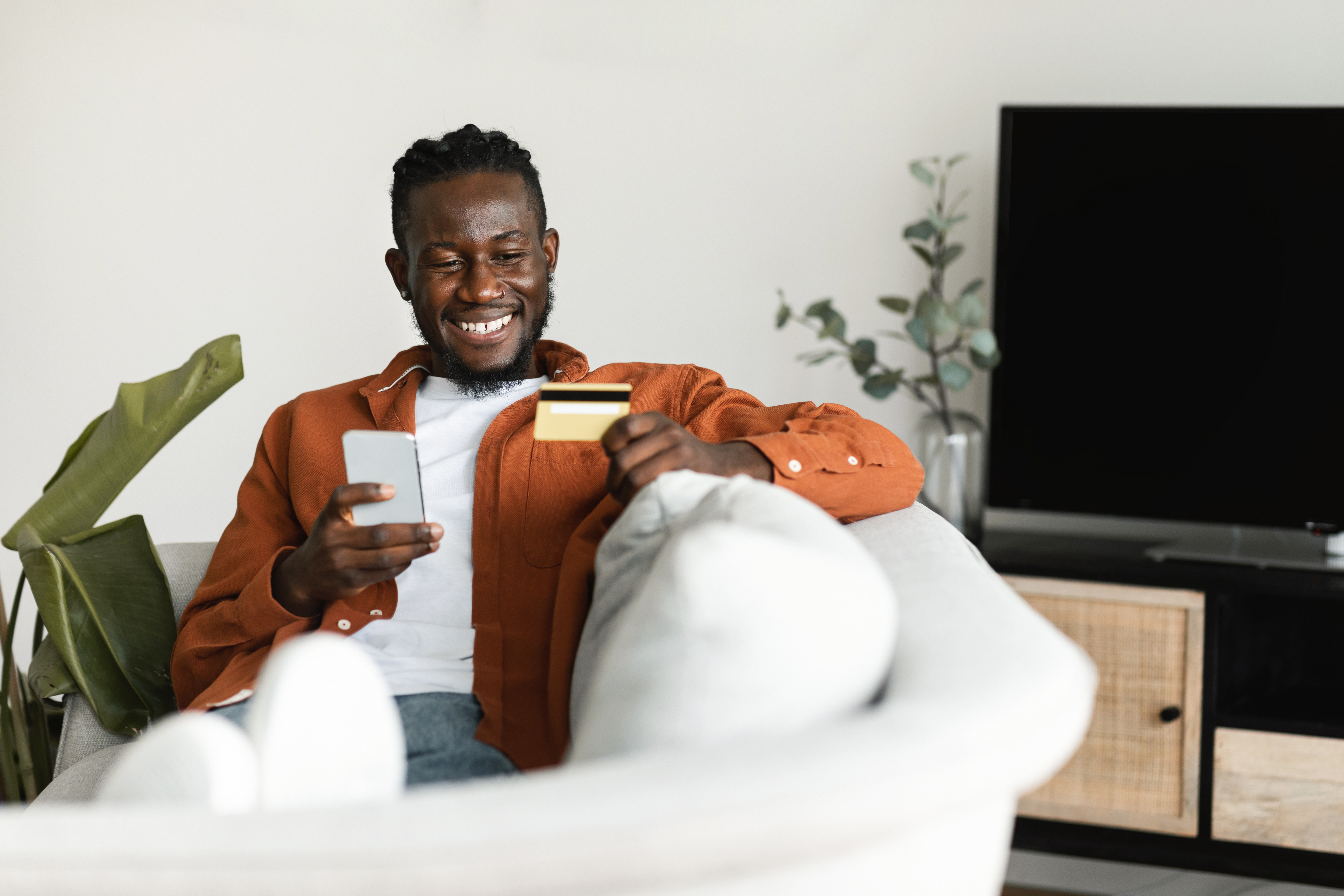 Man Holding Debit Credit Card in Hand and Using Smartphone, Making Financial Transaction While Resting on Couch at Home Man Holding Debit Credit Card in Hand and Using Smartphone, Making Financial Transaction While Resting on Couch at Home